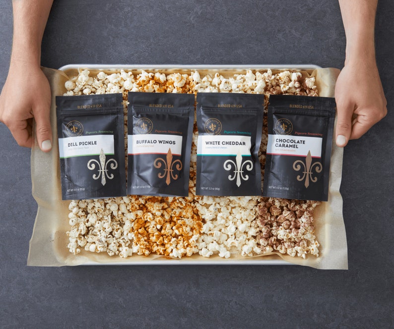 Hands holding backing tray that has 4 seasoning pouches laying on top of seasoned popcorn. Dell Cove Spices