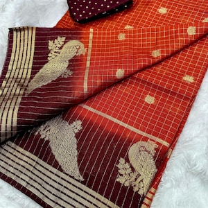 May include: An orange and burgundy saree with gold accents. The saree features a grid pattern and gold peacock designs. A matching burgundy blouse piece with gold polka dots is included.