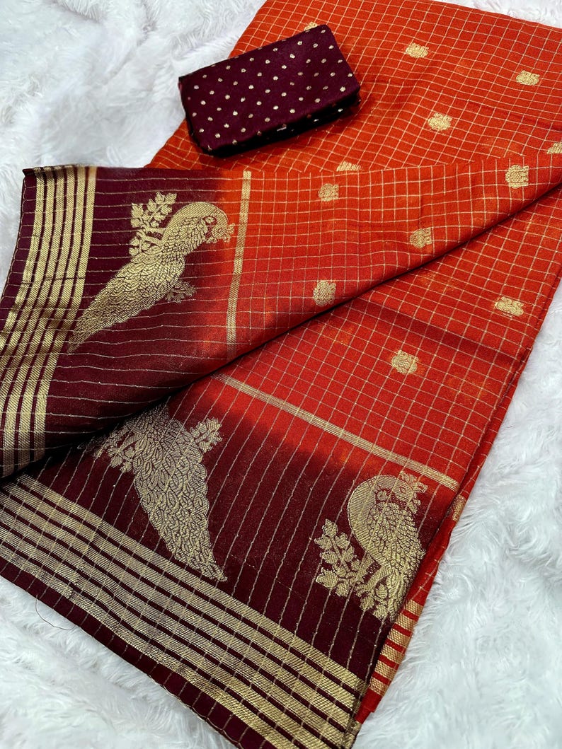 May include: An orange and burgundy saree with gold accents. The saree features a grid pattern and gold peacock designs. A matching burgundy blouse piece with gold polka dots is included.