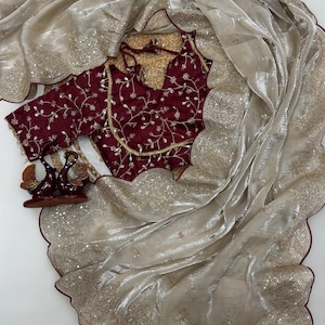 May include: A cream-coloured saree with a scalloped edge, embellished with sequins. A maroon blouse with floral embroidery and gold trim is visible. The saree is draped over the blouse, creating a layered effect. Decorative items are placed near the blouse.