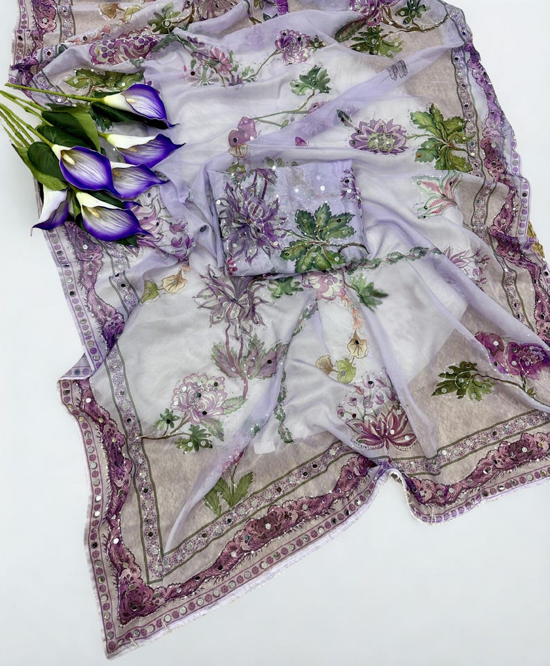 Op de afbeelding: Een doorschijnende, lichtpaarse stof met een bloemenpatroon in tinten paars, groen en wit. De stof is versierd met pailletten en een decoratieve rand. Er is ook een kleine bijpassende vierkante stof aanwezig. Een boeket paarse calla lelies staat links.