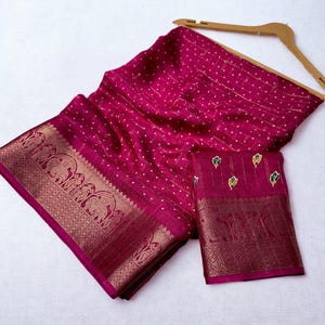 Op de afbeelding: Een magenta sari met een gouden rand en een ingewikkeld olifantenontwerp. De stof heeft een stippenpatroon en is over een houten kleerhanger gedrapeerd. Er is ook een bijpassend stuk stof met een vergelijkbare rand aanwezig.