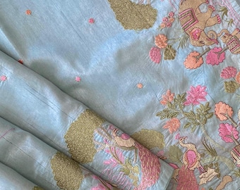 May include: A light blue textile with intricate embroidery. The design features pink, peach, and gold floral and animal motifs, including elephants and peacocks. The fabric is folded, showcasing the detailed patterns.