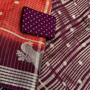 May include: A vibrant textile display featuring a red and burgundy saree with silver accents and a peacock design. A small, dark purple fabric with white polka dots is placed on top. The saree has a checkered pattern.