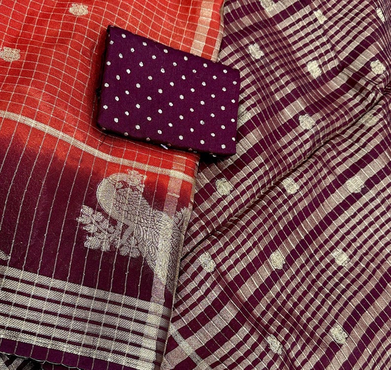 May include: A vibrant textile display featuring a red and burgundy saree with silver accents and a peacock design. A small, dark purple fabric with white polka dots is placed on top. The saree has a checkered pattern.
