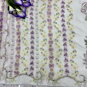 Op de afbeelding: Een lichtpaarse stof met een bloemenpatroon in paarse, gele en groene tinten. De stof heeft een decoratieve rand met kleine spiegelaccenten. Een boeket paarse calla lelies ligt op de stof.