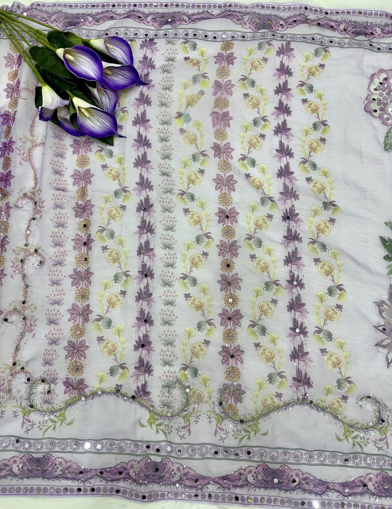 Op de afbeelding: Een lichtpaarse stof met een bloemenpatroon in paarse, gele en groene tinten. De stof heeft een decoratieve rand met kleine spiegelaccenten. Een boeket paarse calla lelies ligt op de stof.