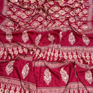 May include: A deep magenta fabric featuring a repeating pattern of cream and gold floral motifs interspersed with small reflective circular mirrors. The lower section displays a border of teardrop-shaped designs and cream-colored fabric tassels.