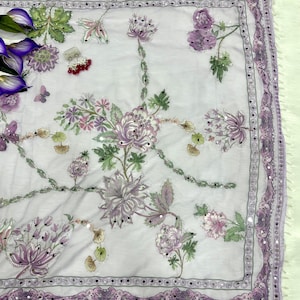Op de afbeelding: Een witte sjaal met een bloemmotief in tinten paars, roze en groen. De sjaal heeft een decoratieve rand met kleine spiegelaccenten. Het bloemmotief bevat gedetailleerd borduurwerk en kralenwerk, wat een delicate en sierlijke uitstraling cre&euml;ert.