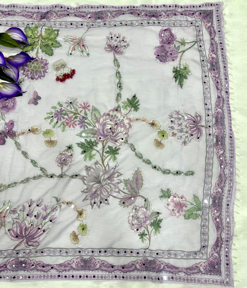 Op de afbeelding: Een witte sjaal met een bloemmotief in tinten paars, roze en groen. De sjaal heeft een decoratieve rand met kleine spiegelaccenten. Het bloemmotief bevat gedetailleerd borduurwerk en kralenwerk, wat een delicate en sierlijke uitstraling cre&euml;ert.