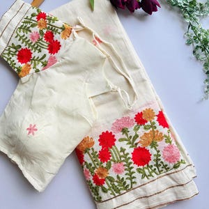 May include: Cream-coloured saree and blouse set. The blouse and saree border are adorned with floral embroidery in red, pink, and orange. The set is made of a lightweight fabric, and the blouse has tie closures.