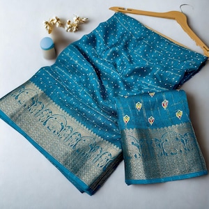 Op de afbeelding: Een turquoise sari met een gouden rand met olifantenmotieven. De stof heeft een stippenpatroon en is over een houten kleerhanger gedrapeerd. Een bijpassend stuk stof is eronder gevouwen. Decoratieve elementen zijn zichtbaar.