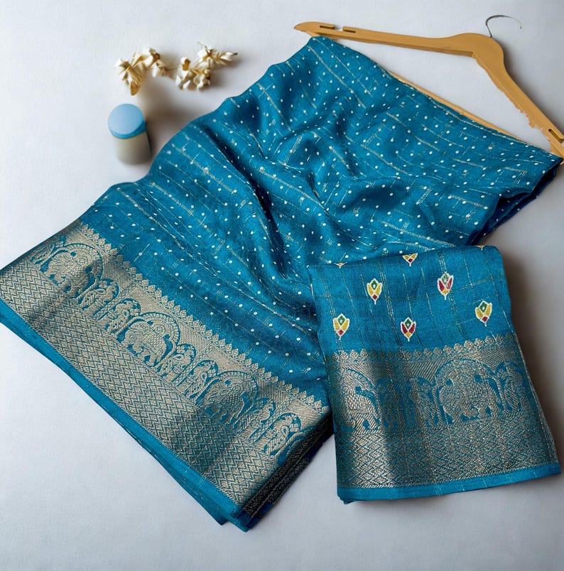 Op de afbeelding: Een turquoise sari met een gouden rand met olifantenmotieven. De stof heeft een stippenpatroon en is over een houten kleerhanger gedrapeerd. Een bijpassend stuk stof is eronder gevouwen. Decoratieve elementen zijn zichtbaar.