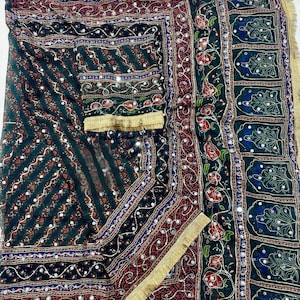 May include: A dark green fabric textile featuring intricate embroidery with red floral patterns, gold metallic thread accents, and small reflective mirror embellishments. The edges are finished with a gold fringe trim, creating a decorative border.