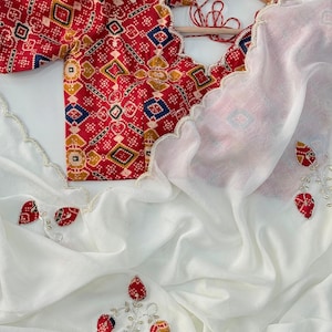 May include: A white saree with a red and patterned blouse. The blouse has a scalloped neckline and short sleeves. The saree is decorated with red and white floral embroidery. The blouse is hanging on a wooden hanger.