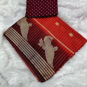 May include: A red and maroon saree with gold accents and bird motifs. The saree has a checkered pattern and striped border. A maroon blouse piece with gold polka dots is included.