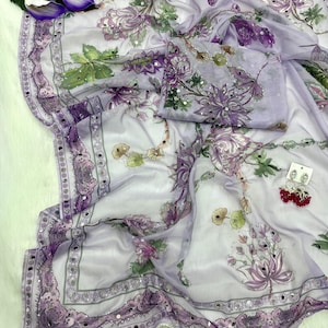Op de afbeelding: Een lichtpaarse bloemen sari met een bijpassend blouse-stuk. De sari heeft een gedetailleerde rand met kleine spiegels en een bloemenpatroon in paarse, groene en witte tinten. Een paar rode oorbellen is ook zichtbaar.