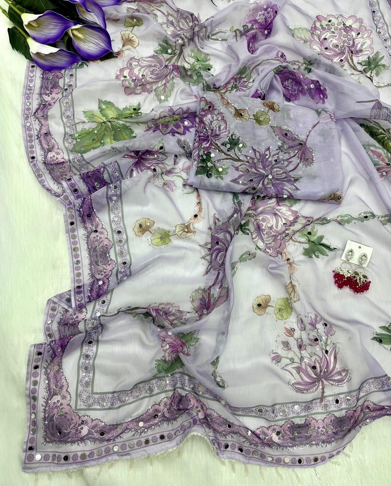 Op de afbeelding: Een lichtpaarse bloemen sari met een bijpassend blouse-stuk. De sari heeft een gedetailleerde rand met kleine spiegels en een bloemenpatroon in paarse, groene en witte tinten. Een paar rode oorbellen is ook zichtbaar.
