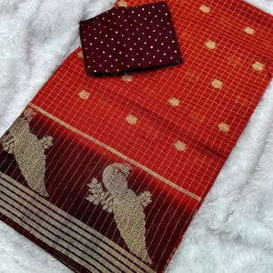 May include: A red and gold saree with a checkered pattern and a maroon blouse. The saree features a decorative border with gold bird motifs. The blouse is maroon with gold polka dots. The saree is displayed on a white, fluffy surface.