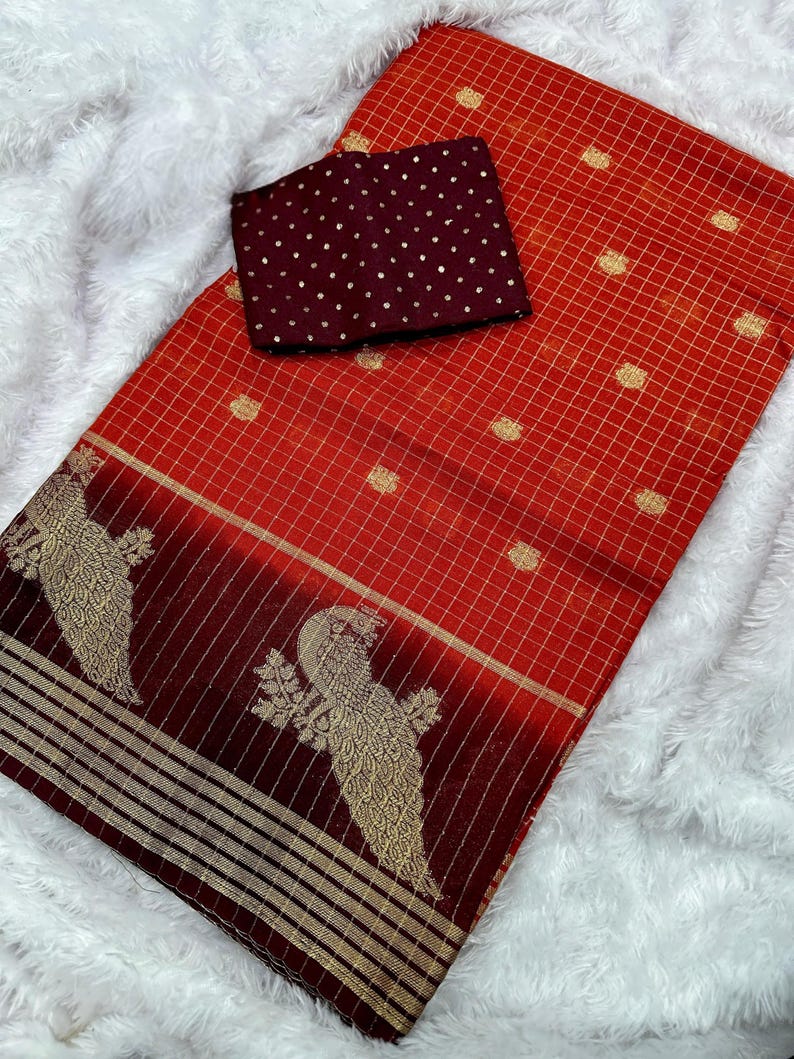 May include: A red and gold saree with a checkered pattern and a maroon blouse. The saree features a decorative border with gold bird motifs. The blouse is maroon with gold polka dots. The saree is displayed on a white, fluffy surface.