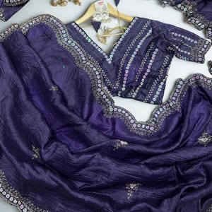 Puede incluir: Un conjunto de sari y blusa de color púrpura oscuro. La blusa tiene cuello en V y está adornada con lentejuelas y bordados. El sari presenta un borde detallado con lentejuelas y bordados. La tela tiene una apariencia texturizada.