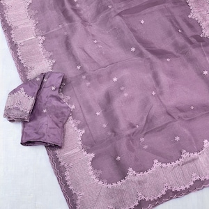Könnte beinhalten: Ein malvenfarbener Sari aus transparentem, leichtem Stoff mit verstreuten kleinen Blumenstickereien. Der Rand ist mit einer aufwendigen, bogenförmigen Spitzenverzierung in einem helleren Malventon versehen, dazu ein passendes Kurzarm-Oberteil.