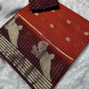 May include: A vibrant orange and burgundy saree with gold accents and a matching blouse. The saree features a grid pattern and peacock motifs. The blouse is a solid burgundy with gold polka dots. The fabric appears to be silk.