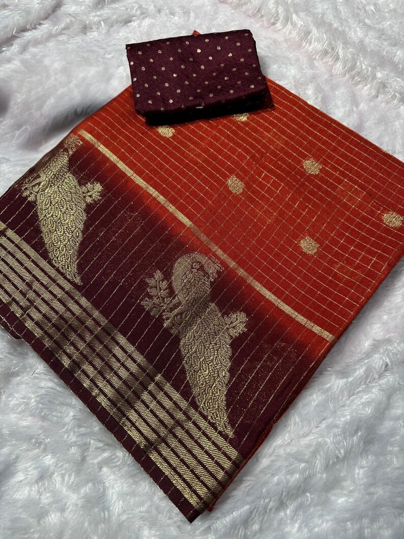 May include: A vibrant orange and burgundy saree with gold accents and a matching blouse. The saree features a grid pattern and peacock motifs. The blouse is a solid burgundy with gold polka dots. The fabric appears to be silk.
