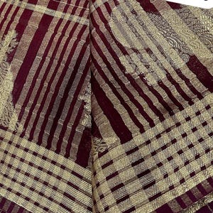 May include: Close-up of a textile with a pattern of alternating burgundy and gold stripes and checks. The fabric has a shimmering quality, with the gold elements catching the light. The overall design suggests a decorative or formal textile.