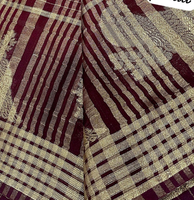 May include: Close-up of a textile with a pattern of alternating burgundy and gold stripes and checks. The fabric has a shimmering quality, with the gold elements catching the light. The overall design suggests a decorative or formal textile.