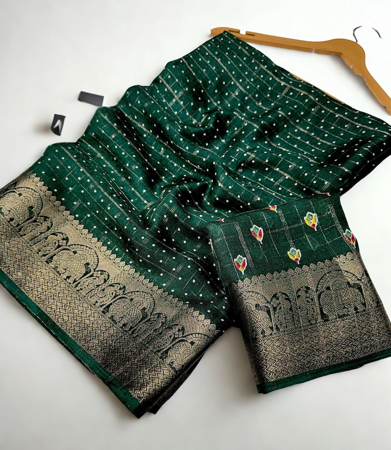 Op de afbeelding: Een donkergroen textiel met een gouden rand en een herhalend patroon van olifanten. De stof heeft een gestippeld en gestreept ontwerp. Het textiel is over een houten hanger gedrapeerd.