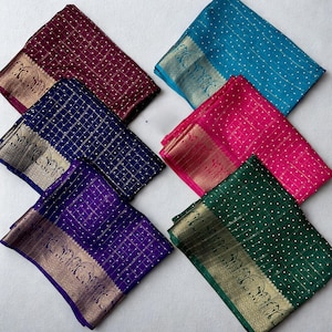 Op de afbeelding: Een assortiment gevouwen zijden sari's in verschillende kleuren. Elke sari heeft een gedetailleerde gouden rand met patronen en kleine witte stippen. De kleuren zijn onder meer turquoise, roze, paars, marineblauw, bordeauxrood en groen.