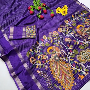 May include: A vibrant purple saree with a detailed peacock and floral design. The saree features a matching purple blouse piece and a decorative border. The fabric has a subtle striped texture. A green and red decorative item and yellow flowers are placed on top.