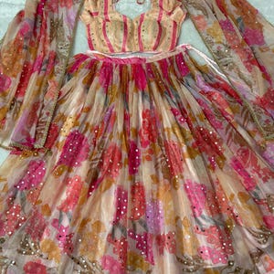Puede incluir: Un conjunto de tres piezas que incluye una blusa sin mangas con rayas verticales en color melocotón y rosa, una falda plisada a juego con un estampado floral vibrante en tonos rosa, naranja y amarillo, y una dupatta transparente con bordados dorados.