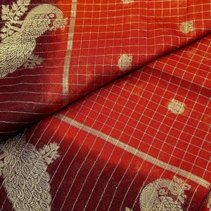 May include: Close-up of a red fabric with a gold grid pattern and peacock design. The fabric has a gradient effect, transitioning from a deep burgundy to a vibrant red. The peacock design is detailed with gold thread.