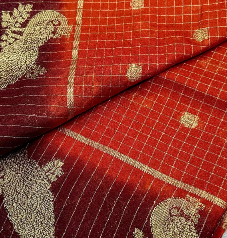 May include: Close-up of a red fabric with a gold grid pattern and peacock design. The fabric has a gradient effect, transitioning from a deep burgundy to a vibrant red. The peacock design is detailed with gold thread.