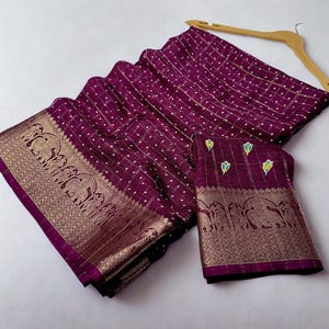 Op de afbeelding: Een pruimkleurige sari met een gouden rand met olifantenmotieven. De sari heeft een rasterpatroon met witte stippen en is over een houten kleerhanger gedrapeerd. Er is ook een bijpassend stuk stof aanwezig.