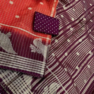 May include: A vibrant textile featuring a combination of red and burgundy hues. The fabric showcases a grid pattern with gold accents and intricate peacock motifs. A small, dark purple pouch with white polka dots is also present.