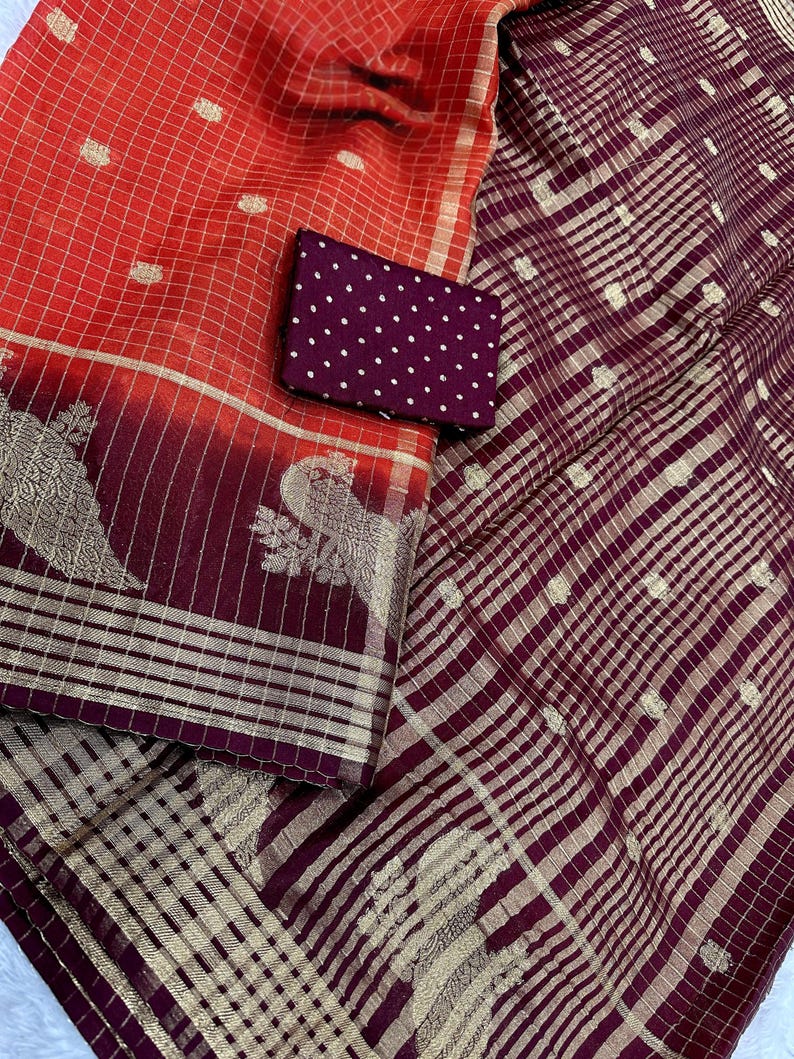 May include: A vibrant textile featuring a combination of red and burgundy hues. The fabric showcases a grid pattern with gold accents and intricate peacock motifs. A small, dark purple pouch with white polka dots is also present.