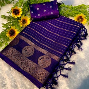 May include: A vibrant purple saree with gold-colored decorative borders and intricate patterns. The saree features a matching purple blouse piece with floral embroidery. The design includes elephant and peacock motifs, and the saree has a fringed edge.