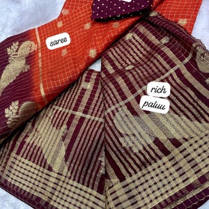 May include: An orange saree with a gold grid pattern and a matching maroon blouse with white polka dots. The saree has a rich maroon pallu with gold stripes and floral designs.