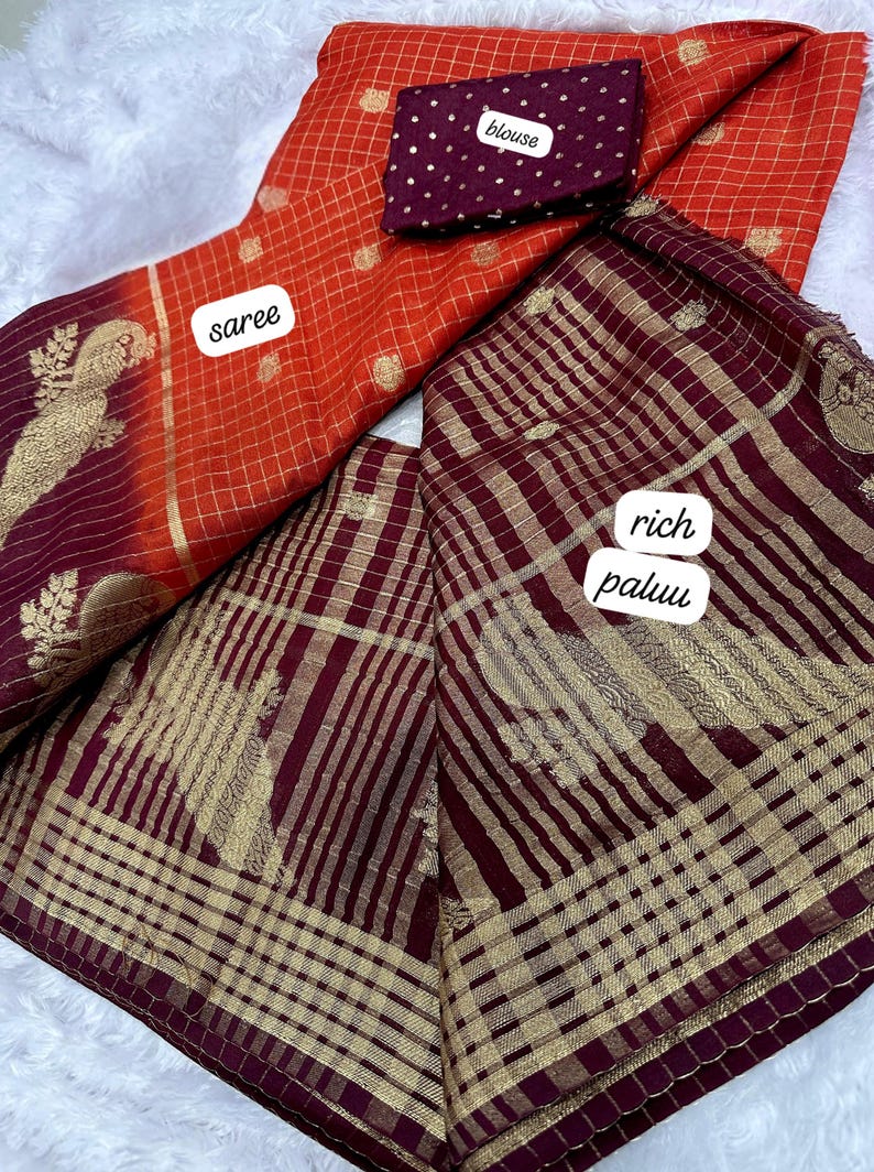 May include: An orange saree with a gold grid pattern and a matching maroon blouse with white polka dots. The saree has a rich maroon pallu with gold stripes and floral designs.