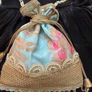 Puede incluir: Un bolso con cordón y estampado floral con adornos dorados y una correa de perlas. El bolso presenta un fondo azul claro con diseños florales rosas y beige. El ribete dorado está adornado con cuentas blancas. La correa es una doble hilera de perlas.