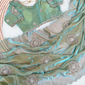 Puede incluir: Un conjunto de sari y blusa verde mar. El sari presenta intrincados patrones florales bordados y un borde festoneado. La blusa tiene bordados detallados en las mangas y el escote. La tela tiene una calidad brillante e iridiscente.