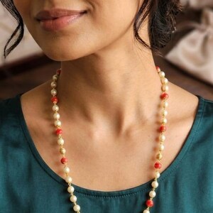 Puede incluir: Un collar largo que presenta un patrón repetitivo de perlas blancas redondas, cuentas rojas vibrantes y pequeños espaciadores de metal dorado con textura. Las cuentas están ensartadas en una fina cadena dorada sobre una prenda verde.