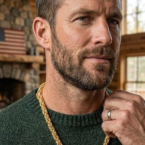 Puede incluir: Una cadena gruesa de color dorado con un patrón de eslabones entrelazados de estilo bizantino. El collar se lleva alrededor del cuello, sobre un suéter de punto texturizado de color verde oscuro. La cadena tiene un acabado pulido y un cierre.