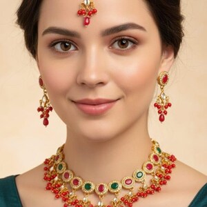 Puede incluir: Juego de joyas doradas con collar, pendientes y tocado. El collar y los pendientes tienen detalles de piedras preciosas rojas y verdes. El tocado tiene una gema roja central y cuentas rojas colgantes.