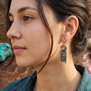 Puede incluir: Un pendiente colgante de plata con un diseño de pájaro estilizado, que presenta intrincados patrones geométricos y detalles en negro oxidado. El pendiente cuelga de un fino gancho de plata y tiene una forma rectangular con bordes redondeados.