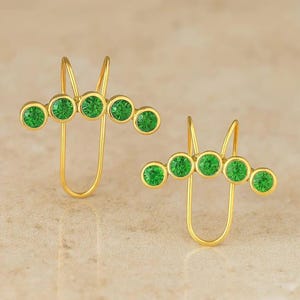 Puede incluir: Un par de pendientes ear cuff dorados, cada uno con una fila curva de cinco piedras redondas de color verde vibrante engastadas en metal. El diseño incluye una estructura de alambre en forma de U que se extiende hacia arriba desde la base.