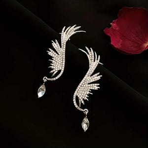 Puede incluir: Un par de pendientes tipo ear cuff plateados con forma de alas, incrustados con pequeños cristales transparentes. Cada ala presenta una gota de cristal transparente de talla marquesa colgando en la parte inferior. Diseño simétrico y detallado.